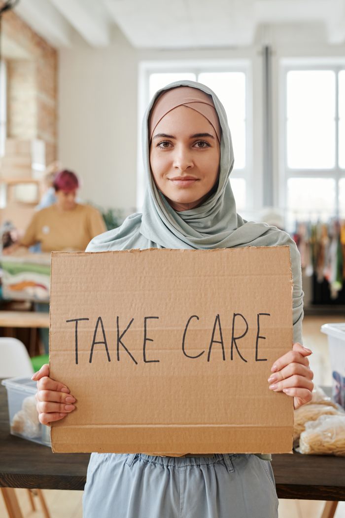 Young woman in hijab proudly displaying a 'Take Care' sign indoors.
